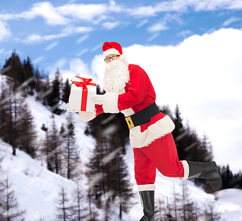 christmas, holidays and people concept - man in costume of santa claus running with gift box over snowy mountains background