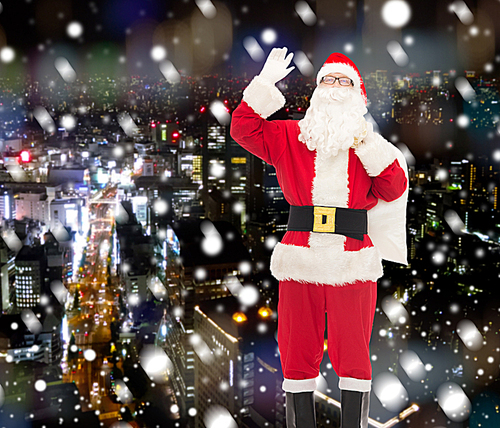 christmas, holidays, gesture and people concept - man in costume of santa claus with bag waving hand over snowy night city background