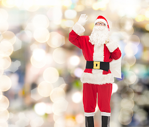 christmas, holidays, gesture and people concept - man in costume of santa claus with bag waving hand over lights background