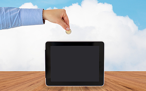 business, finance, investment, money saving and e-commerce concept - close up of hand putting coin into into tablet pc computer with black blank screen over blue sky and wooden floor background