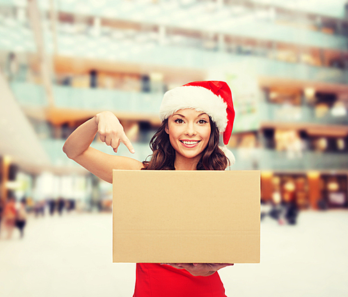 christmas, winter, holidays, delivery and people concept - smiling woman in santa helper hat with parcel box over shopping center background