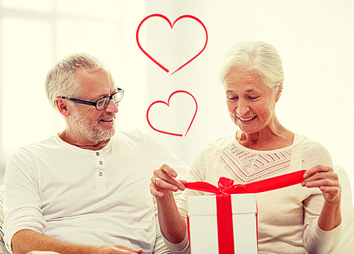 family, holidays, christmas, age and people concept - happy senior couple opening gift box over white room background with red heart shapes