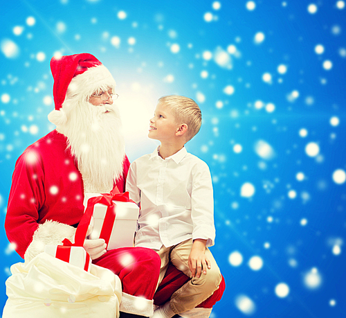 holidays, christmas, childhood and people concept - smiling little boy with santa claus and gifts over blue snowy background