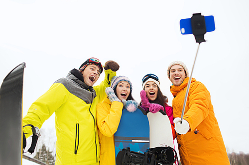 winter sport, leisure, friendship, technology and people concept - happy friends with snowboards and taking picture by smartphone on selfie stick