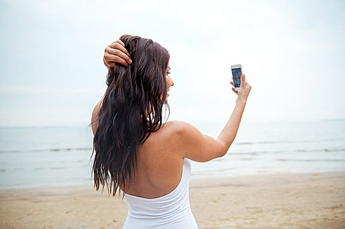 summer, travel, technology and people concept -  young woman taking selfie with smartphone on beach from back