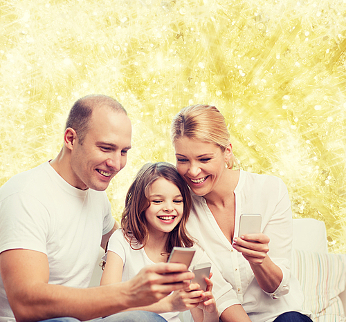 family, holidays, technology and people concept - smiling mother, father and little girl with smartphones over yellow lights background