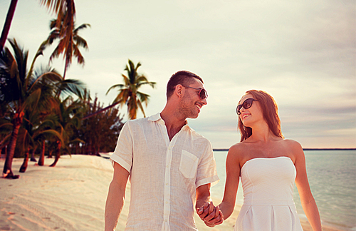 love, people, travel, summer and relations concept - smiling couple wearing sunglasses walking outdoors over tropical beach background