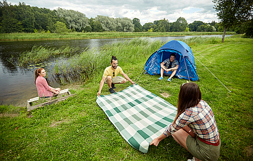 camping, travel, tourism, hike and people concept - happy friends with tent laying picnic blanket at campsite on river bank