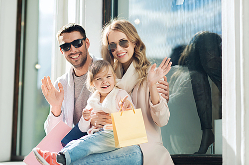 sale, consumerism and people concept - happy family with little child and shopping bags waving hands at shop window in city