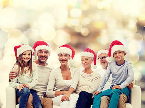 family, happiness, generation, holidays and people concept - happy family in santa helper hats sitting on couch over lights background