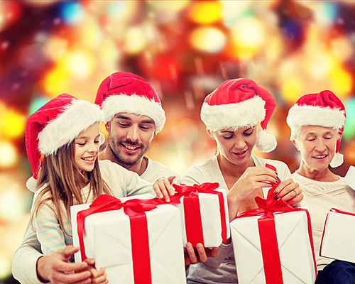 family, happiness, generation, holidays and people concept - happy family in santa helper hats with gift boxes sitting on couch over red lights background