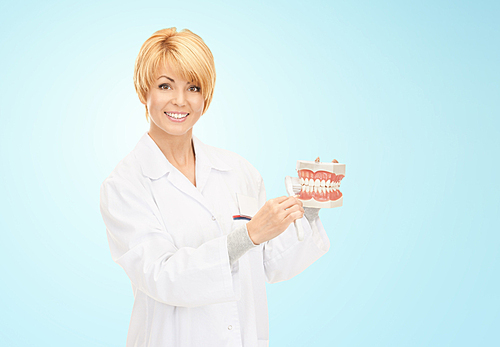 medicine, stomatology, people and dental hygiene concept - smiling female doctor with toothbrush and jaws model teaching how to brush teeth over blue background