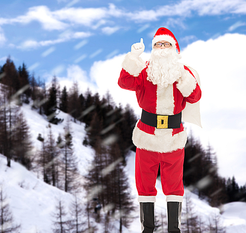 christmas, holidays, gesture and people concept - man in costume of santa claus with bag pointing finger up over snowy mountains background