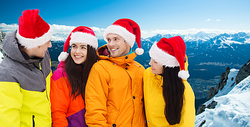 winter holidays, christmas, friendship and people concept - happy friends in santa hats and ski suits talking outdoors over snowy mountains background