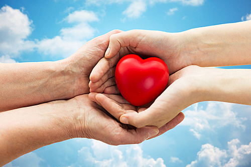 people, age, family, love and health care concept - close up of senior woman and young woman hands holding red heart over blue sky and clouds background