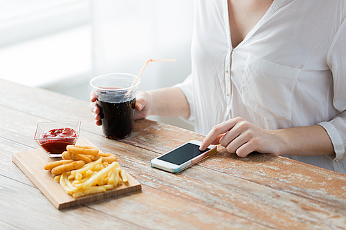 fast food, people, technology and diet concept - close up of woman with smartphone drinking cola and eating french fries, ketchup and deep-fried squid rings at wooden table