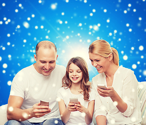 family, christmas holidays, technology and people - smiling mother, father and little girl with smartphones over blue snowy background