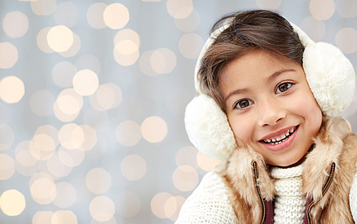 winter, people, christmas happiness concept - happy little girl wearing earmuffs and gloves over holidays lights background