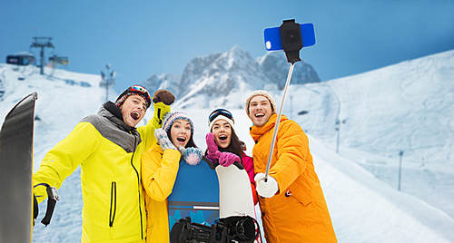 winter sport, leisure, friendship, technology and people concept - happy friends with snowboards and taking picture by smartphone on selfie stick over downhill skiing and mountains background