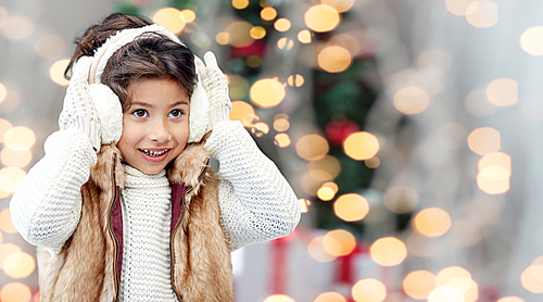 winter, people, childhood and happiness concept - happy little girl wearing earmuffs over christmas tree lights background