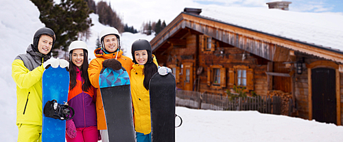 winter, leisure, extreme sport, friendship and people concept - happy friends in helmets with snowboards outdoors over wooden country house background and snow