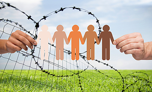 crime, imprisonment, refugee and humanity concept - multiracial couple hands holding chain of paper people pictogram over sky, grass and barb wire background