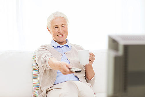 technology, television, age and people concept - happy senior woman watching tv, drinking tea and changing channels by remote control at home