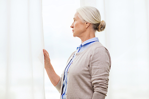 age, loneliness and people concept - lonely senior woman looking through window at home