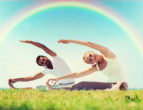 fitness, sport, yoga and people concept - happy couple stretching on mats over rainbow in blue sky background