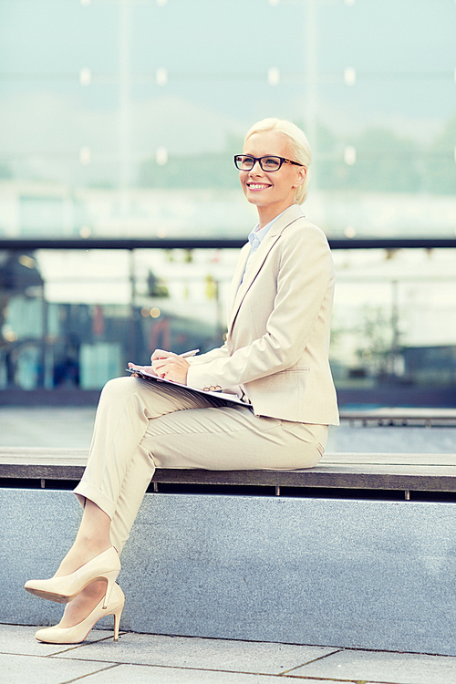 business, people and education concept - young smiling businesswoman in glasses with notepad over office building