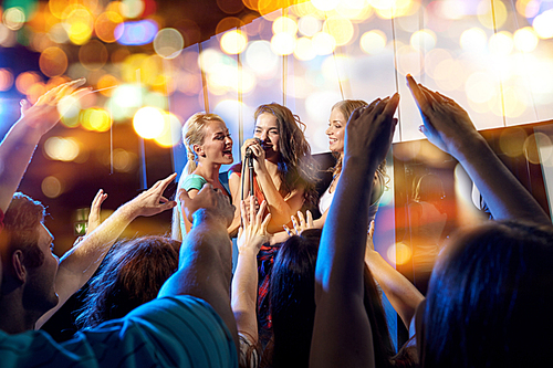 party, holidays, celebration, nightlife and people concept - happy young women singing karaoke in night club behind crowd of music fan