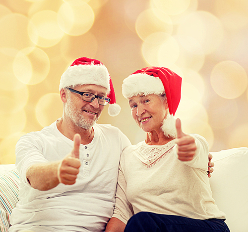 family, holidays, christmas, age and people concept - happy senior couple in santa helper hats sitting on sofa over beige lights background and showing thumbs up gesture