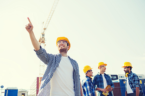 business, building, teamwork and people concept - group of smiling builders in hardhats pointing finger outdoors