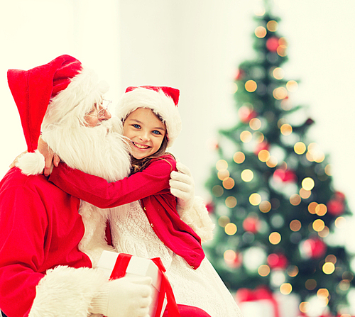 holidays, celebration, childhood and people concept - smiling little girl hugging with santa claus over christmas tree lights background