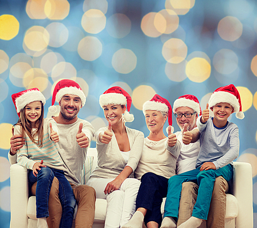 family, happiness, generation, holidays and people concept - happy family in santa helper hats sitting on couch and showing thumbs up gesture over blue lights background