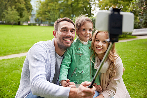 family, childhood, technology and people concept - happy father, mother and little daughter taking picture by smartphone selfie stick in park