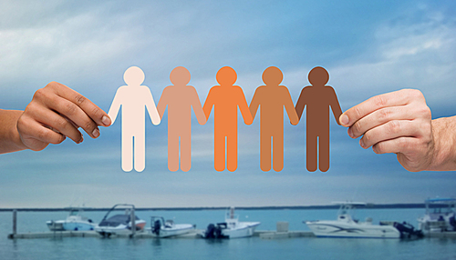 immigration, unity, population, race and humanity concept - multiracial couple hands holding chain of paper people pictogram over boats in sea background