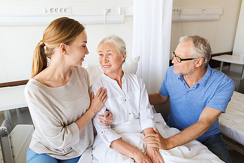 medicine, support, family health care and people concept - happy senior man and young woman visiting and cheering her grandmother lying in bed at hospital ward