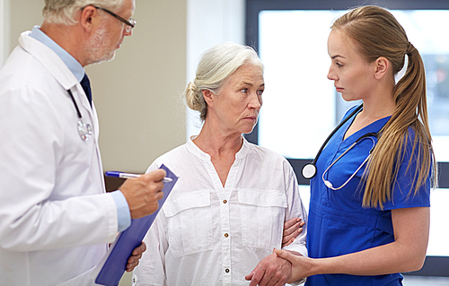 medicine, age, health care and people concept - male doktor with clipboard, young nurse and senior woman patient talking at hospital corridor