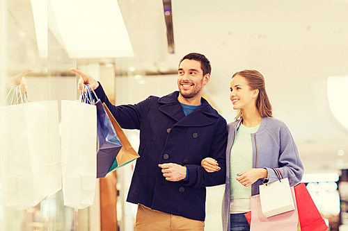 sale, consumerism and people concept - happy young couple with shopping bags pointing finger to shop window in mall