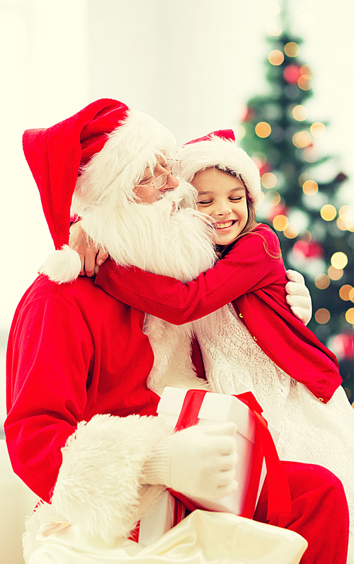 holidays, celebration, childhood and people concept - smiling little girl hugging with santa claus over christmas tree lights background