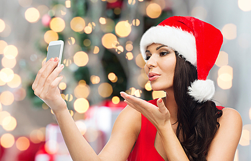 people, holidays, christmas and technology concept - beautiful  woman in red santa hat taking selfie picture by smartphone and sending blow kiss to camera over lights background