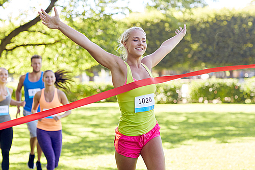 fitness, sport, victory, success and healthy lifestyle concept - happy woman winning race and coming first to finish red ribbon over group of sportsmen running marathon with badge numbers outdoors