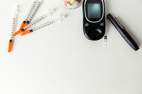 medicine, diabetes, advertisement and health care concept - close up of glucometer with blood sugar test stripe, insulin injection syringes and pills on table