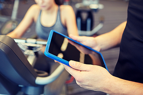 sport, fitness, lifestyle, technology and people concept - close up of trainer hands with tablet pc computer and woman working out on exercise bike in gym