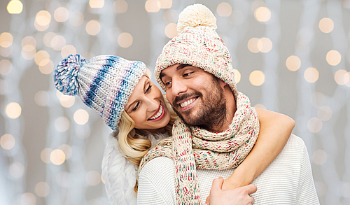 winter, fashion, couple, christmas and people concept - smiling man and woman in hats and scarf hugging over holidays lights background