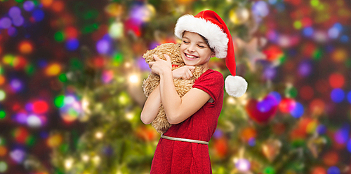 christmas, winter, holidays and childhood concept - smiling girl in santa helper hat with teddy bear over glitter or lights background