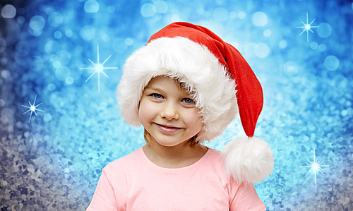 holidays, christmas, childhood and people concept - smiling little girl in santa hat sitting on couch over blue glitter or lights background