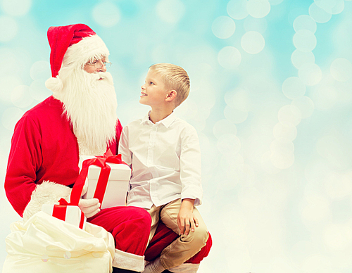 holidays, christmas, childhood and people concept - smiling little boy with santa claus and gifts over blue lights background