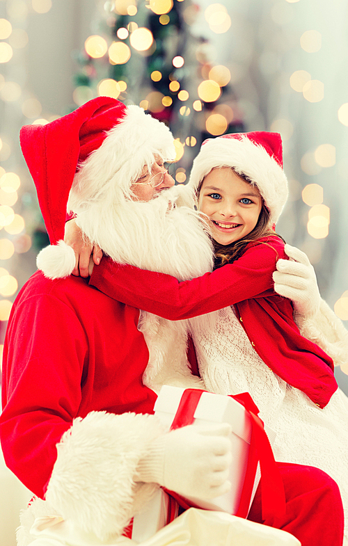 holidays, celebration, childhood and people concept - smiling little girl hugging with santa claus over christmas tree lights background